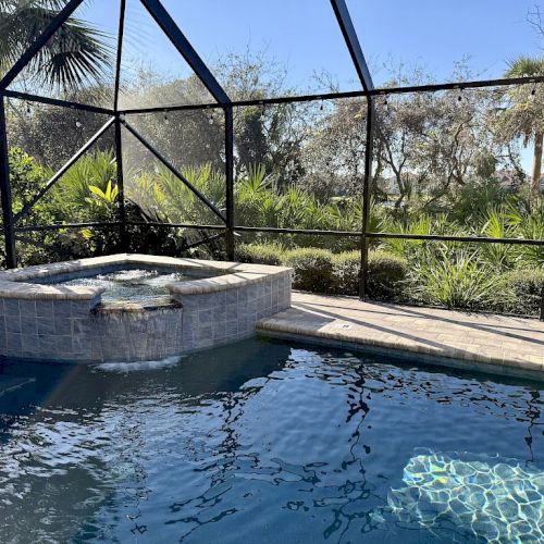 A swimming pool and hot tub are enclosed by a screened structure, surrounded by greenery and trees under a clear blue sky.