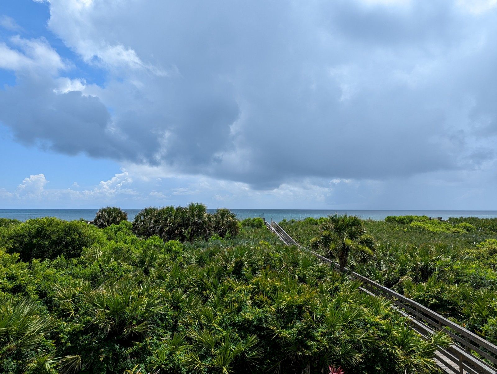 A boardwalk stretches through lush green foliage toward the ocean. Fluffy clouds fill the sky above the tranquil scene.