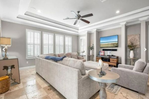 A modern living room with beige sofas, a TV, ceiling fan, window shutters, decorative plants, and tiled flooring.
