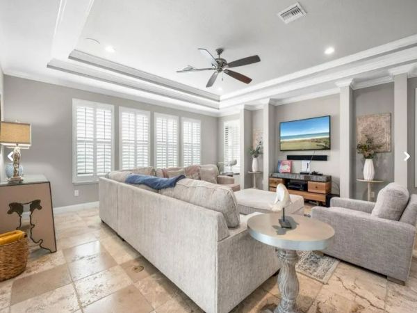 A modern living room with beige sofas, a TV, ceiling fan, window shutters, decorative plants, and tiled flooring.