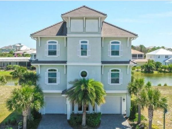 A large three-story house with a tiled roof, surrounded by palm trees, near a body of water.