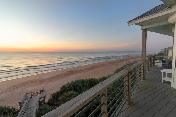 A serene beach scene at sunset, viewed from a wooden deck. The ocean waves gently touch the sandy shore, creating a peaceful atmosphere.