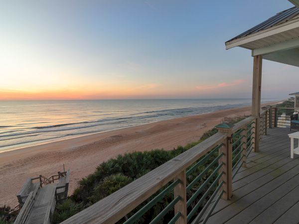 A serene beach scene at sunset, viewed from a wooden deck. The ocean waves gently touch the sandy shore, creating a peaceful atmosphere.