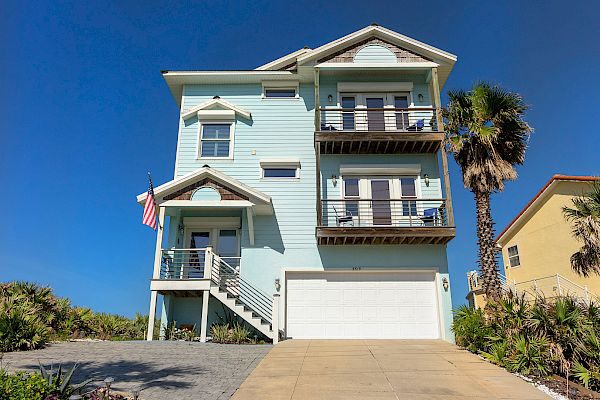 A light blue seaside house with balconies, an American flag, garage, and a palm tree in front under a clear blue sky.