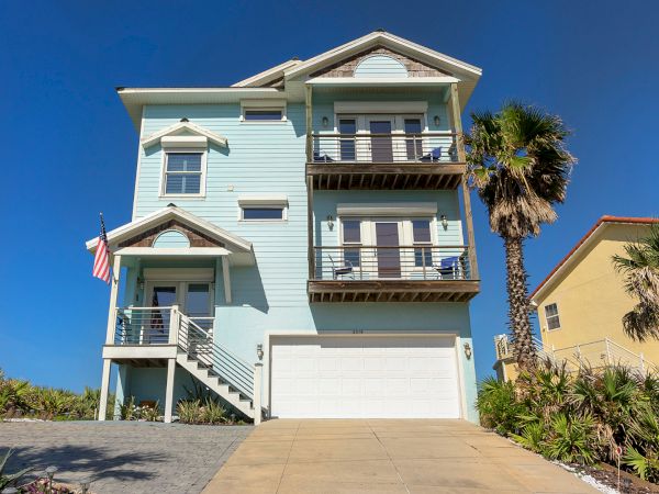 A light blue seaside house with balconies, an American flag, garage, and a palm tree in front under a clear blue sky.