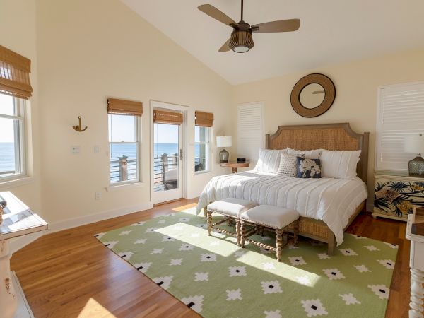 A cozy bedroom with a large bed, sea view through windows, wooden floors, decorative elements, and a light green patterned rug.