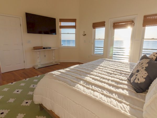 A bright bedroom with ocean views through windows, a bed with white linens, a wall-mounted TV, and a decorative table underneath.