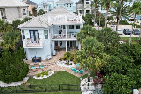 A two-story house with a garden, palm trees, lawn chairs, and nearby parked cars; ocean view in the background completes the scene.