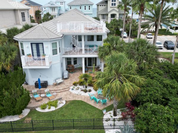 A two-story house with a garden, palm trees, lawn chairs, and nearby parked cars; ocean view in the background completes the scene.