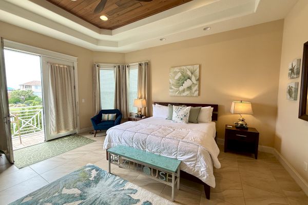 A cozy bedroom with a large bed, blue chair, lamps, artwork, and a view of a balcony with trees and houses outside.