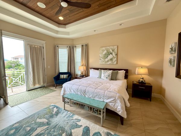 A cozy bedroom with a large bed, blue chair, lamps, artwork, and a view of a balcony with trees and houses outside.