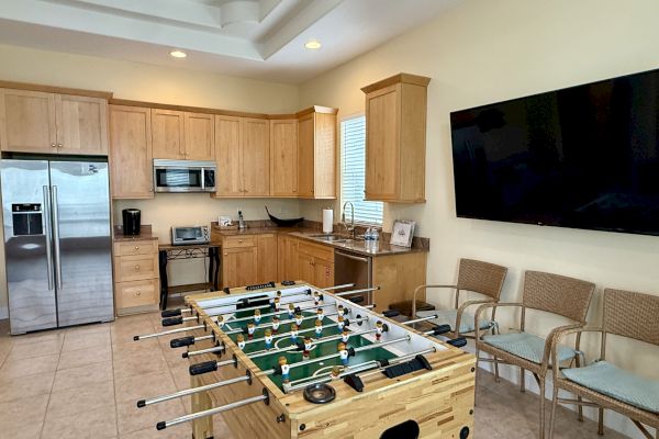 A kitchen with wooden cabinets, stainless steel fridge, microwave, foosball table, wicker chairs, and a large wall-mounted TV.