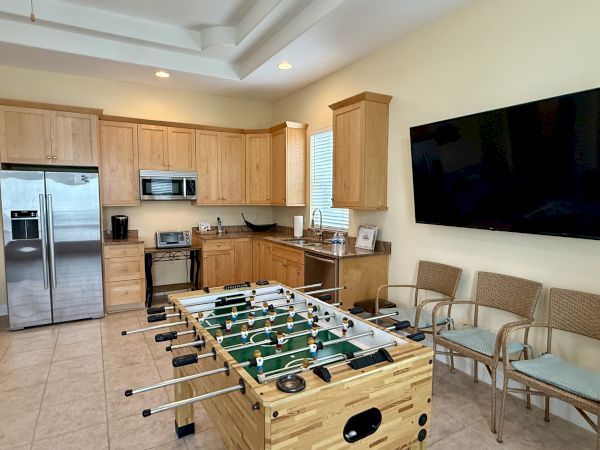 A kitchen with wooden cabinets, stainless steel fridge, microwave, foosball table, wicker chairs, and a large wall-mounted TV.