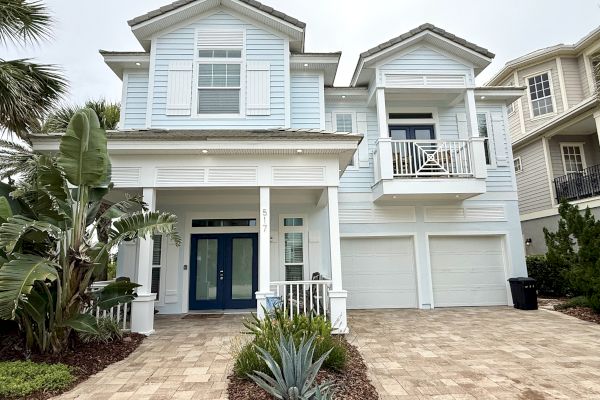 A light blue two-story house with a double garage and balcony, surrounded by tropical plants.