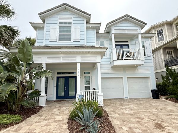 A light blue two-story house with a double garage and balcony, surrounded by tropical plants.