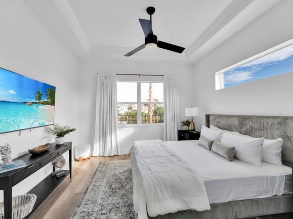 A modern bedroom with a large bed, a wall-mounted TV, a ceiling fan, and a window view, decorated in neutral tones.