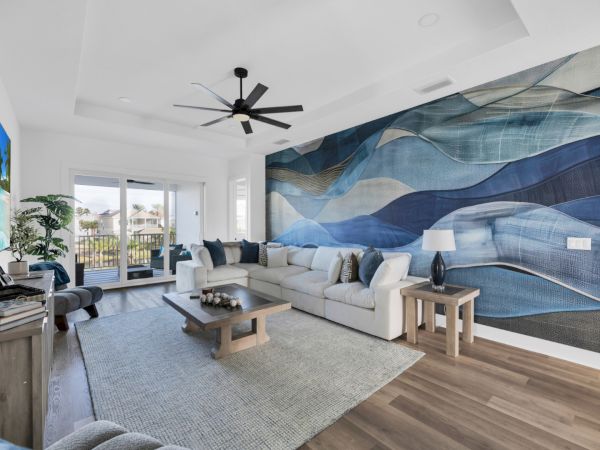 A modern living room with a large white sectional, abstract blue wall art, ceiling fan, TV, and view of a balcony outside the window.