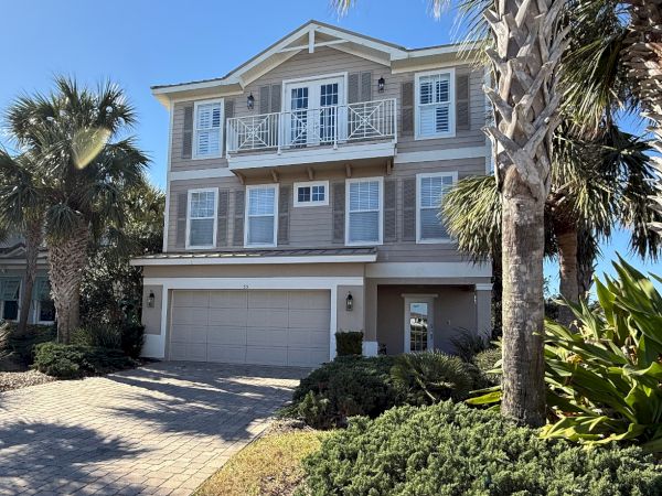 A two-story house with a garage, surrounded by palm trees and landscaped greenery, under a clear blue sky.