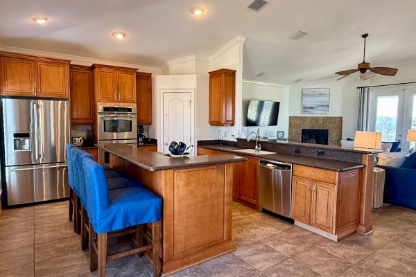 A modern kitchen with wood cabinets, stainless steel appliances, blue barstools, a bar counter, and a view of the living area.
