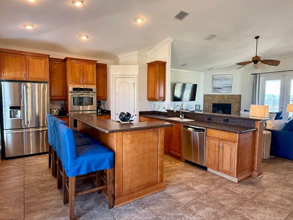 A modern kitchen with wood cabinets, stainless steel appliances, blue barstools, a bar counter, and a view of the living area.