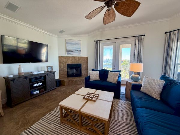 The image shows a modern living room with a blue sofa, wall-mounted TV, and fireplace. A ceiling fan, coffee table, and decor complete the setting.