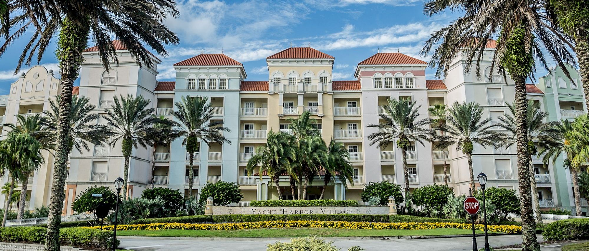 The image shows a large building with red roofs, palm trees, and a landscaped garden under a blue sky with clouds.