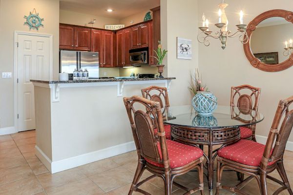 This image shows a cozy kitchen and dining area with wooden cabinets, a round table with red-cushioned chairs, and a decorative chandelier.