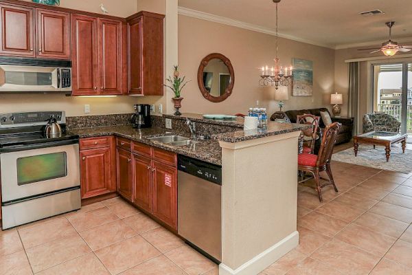 The image shows a kitchen with wooden cabinets, stainless steel appliances, and granite countertops, leading to a dining and living area.
