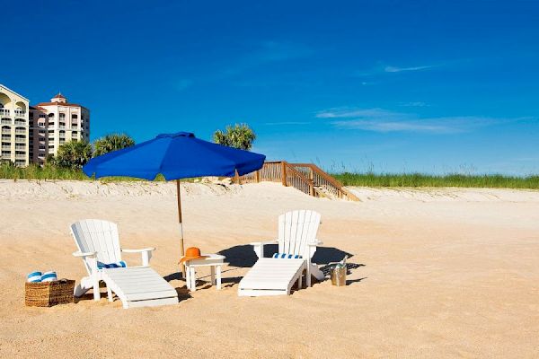 Two white beach chairs, a blue umbrella, and a basket are on a sandy beach with clear skies and some buildings in the background.