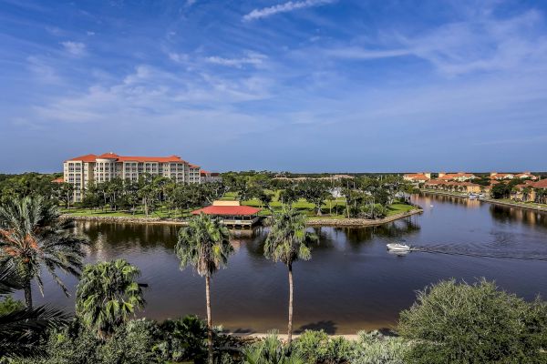 Scenic view of a resort with buildings, palm trees, and a boat on a calm body of water under a clear blue sky.