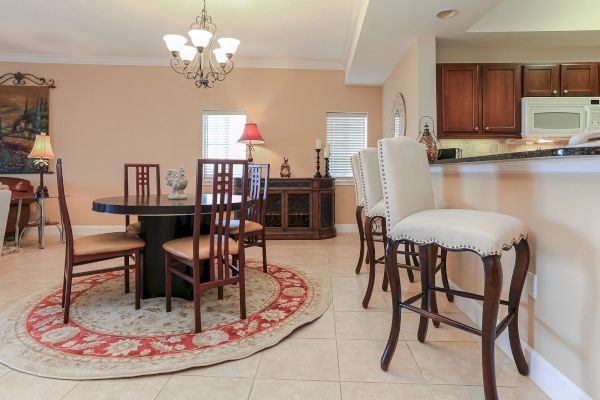 A cozy dining room with a round table, four chairs, bar stools, and warm lighting, featuring a decorative rug and wooden furniture.