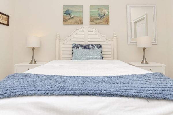 A neatly made bed with blue and white bedding, flanked by lamps on side tables. Beach-themed art and a mirror adorn the walls.