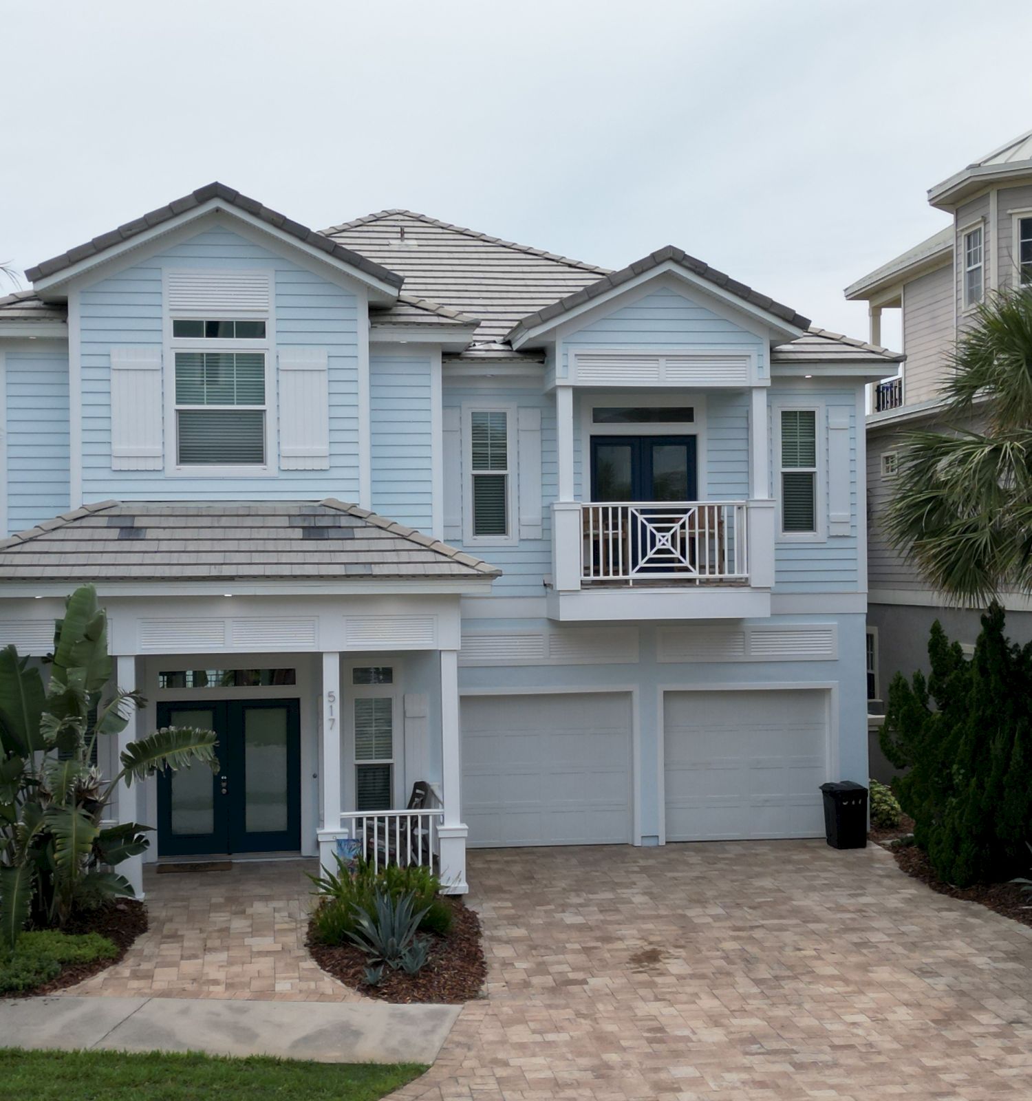 Image of a two-story house with a light blue exterior, surrounded by palm trees and greenery, featuring a paved driveway.