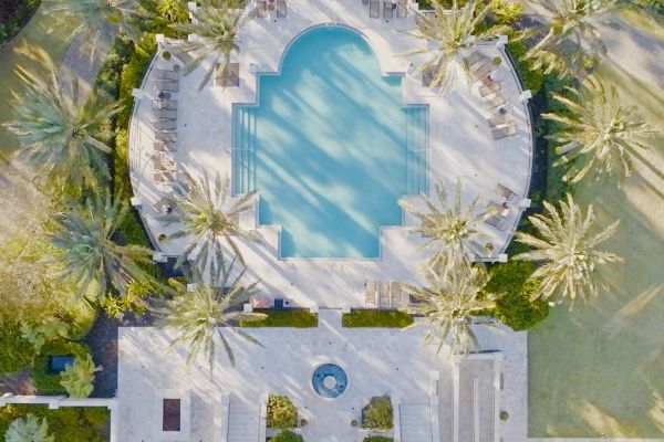 An aerial view of a luxurious pool surrounded by palm trees and lounge chairs, with a small round fountain below, in a sunny setting.