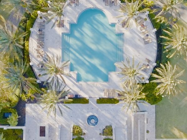 An aerial view of a luxurious pool surrounded by palm trees and lounge chairs, with a small round fountain below, in a sunny setting.