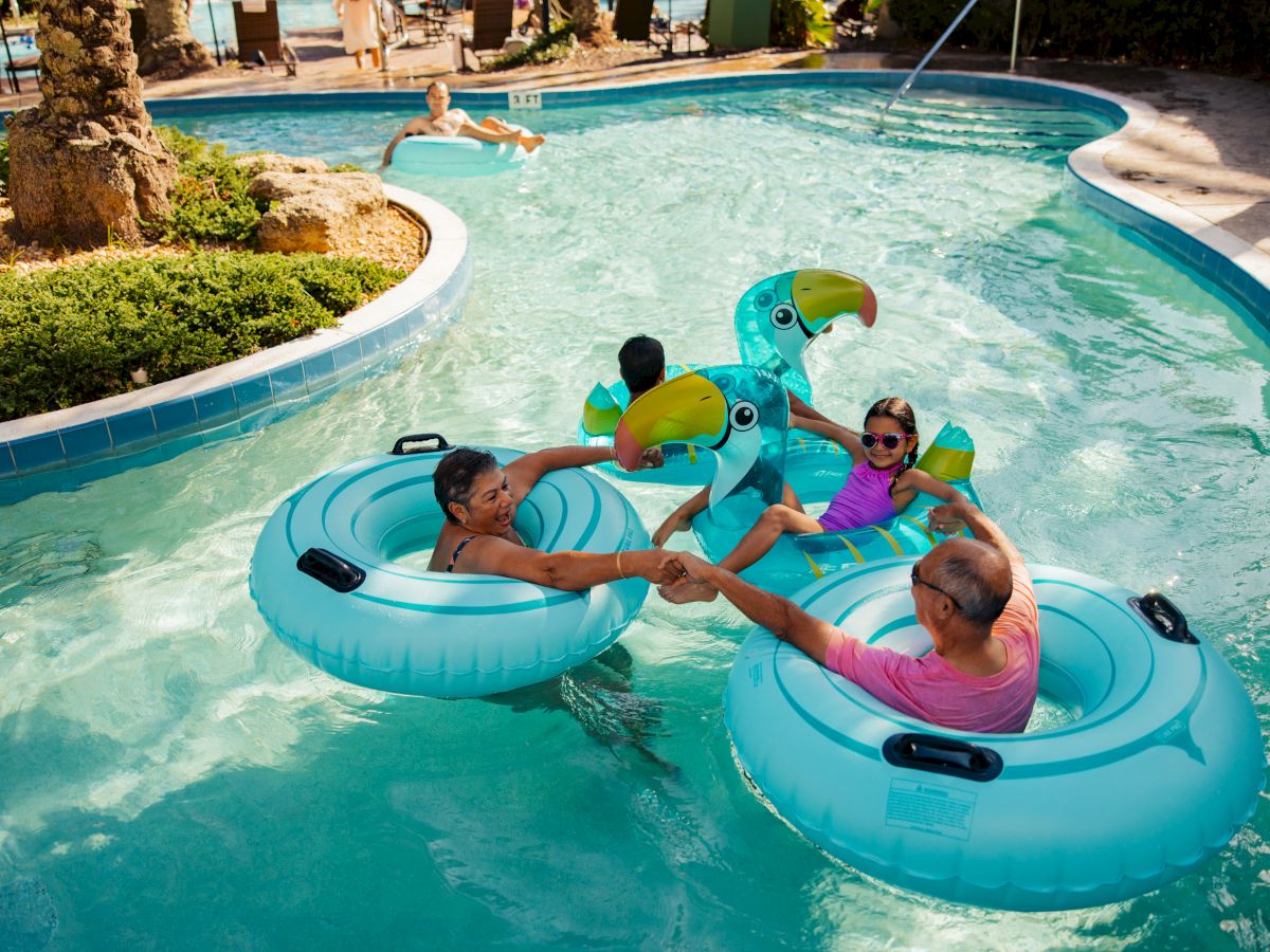 People enjoying a pool on inflatable floats, including a parrot-shaped one, in a lazy river setting under the sun.