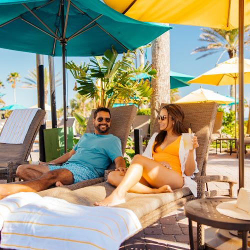 A man and a woman are relaxing on lounge chairs under umbrellas, surrounded by palm trees, on a sunny day.
