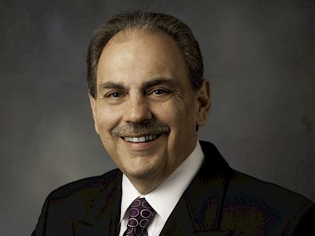 The image shows a person in a suit and tie, smiling, with a dark background.
