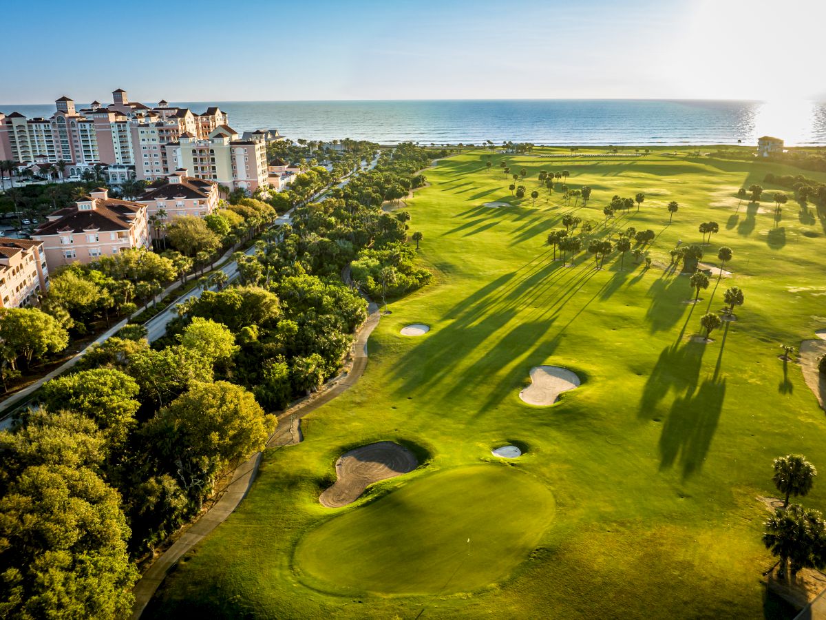 A coastal golf course with sand traps and trees, adjacent to a resort complex, overlooking the sea under a clear blue sky.