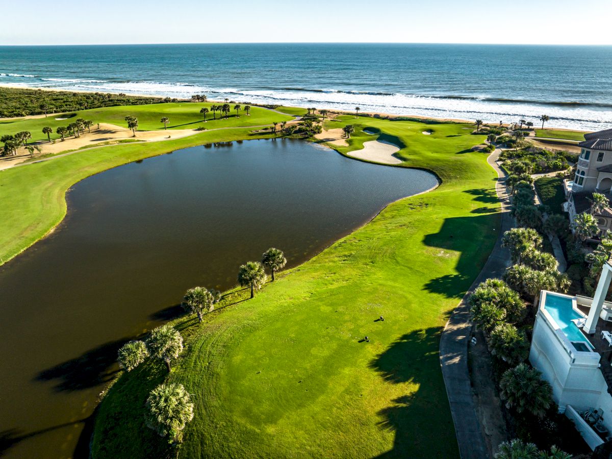 A coastal golf course with lush greens, a sand bunker, palm trees, and a water hazard near the ocean.