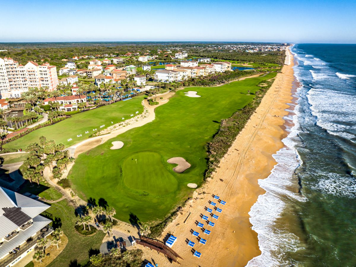 Aerial view of a beachside golf course with buildings in the distance, waves hitting the shoreline, and blue lounge chairs on the sand.
