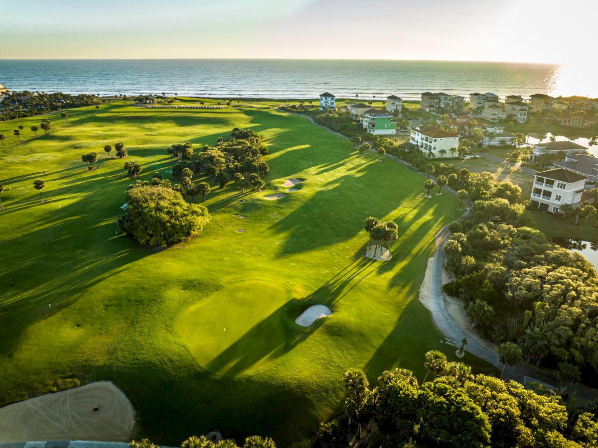 Aerial view of a golf course near the ocean with houses nearby, under a clear sky and sunlight casting long shadows.