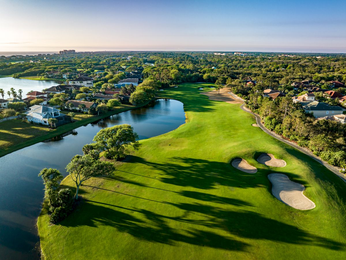 Aerial view of a lush golf course with sand bunkers and water features, surrounded by residential homes and greenery under a clear sky.