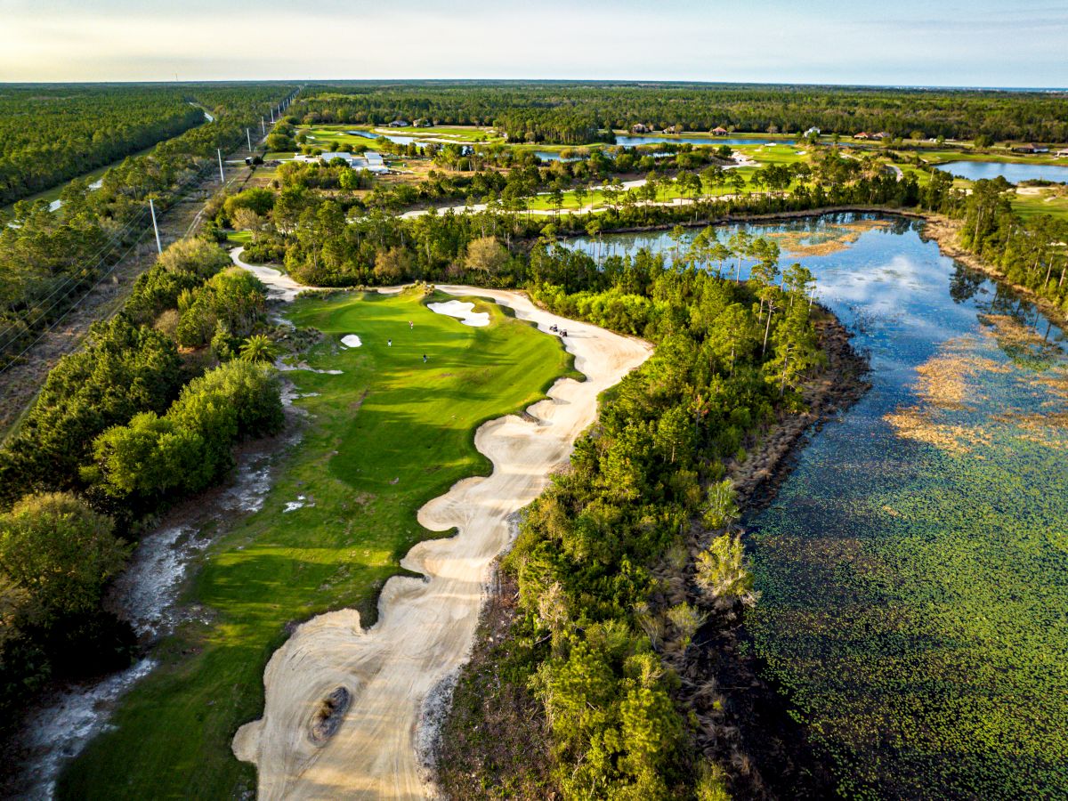 An aerial view of a golf course surrounded by lush greenery and a water body, set against a backdrop of expansive landscapes.