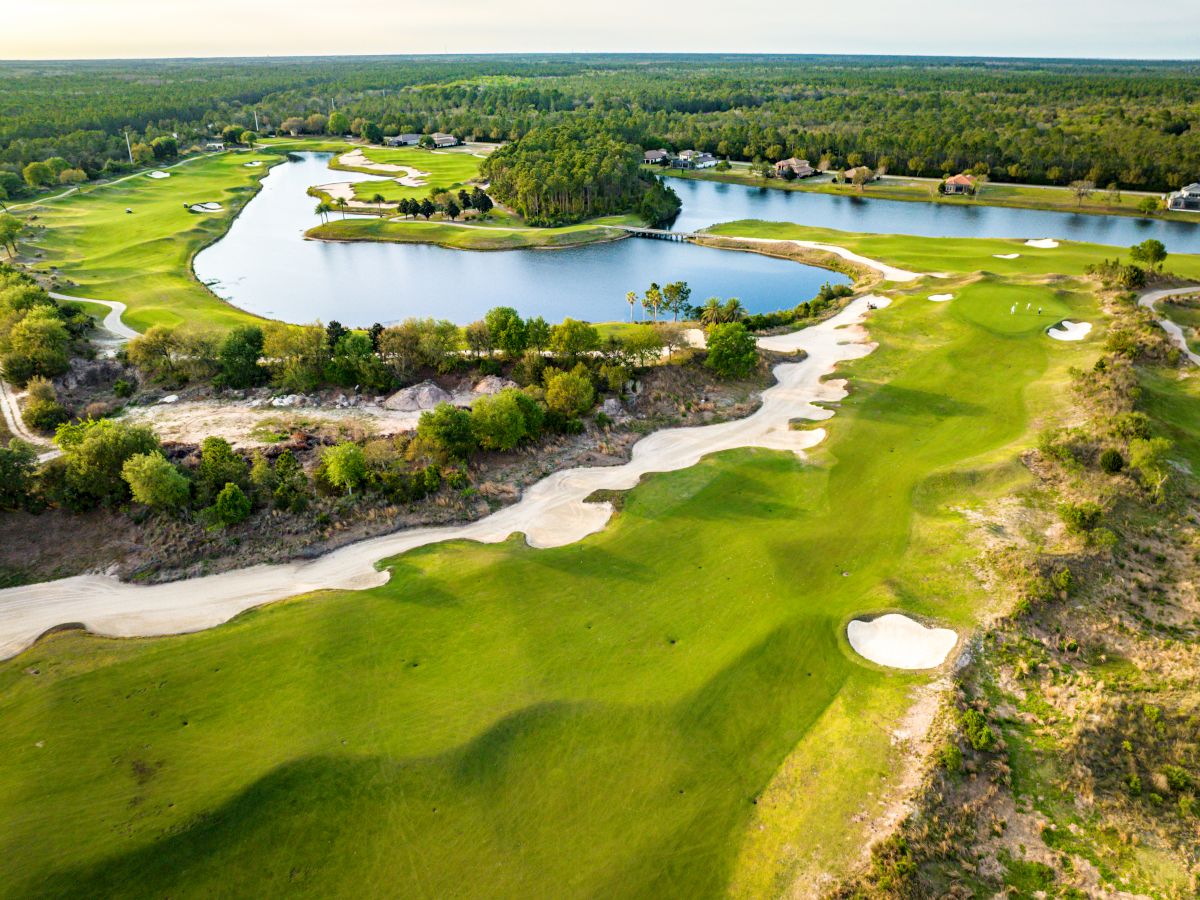 Aerial view of a golf course featuring lush green fairways, sand bunkers, and a lake surrounded by trees and pathways.