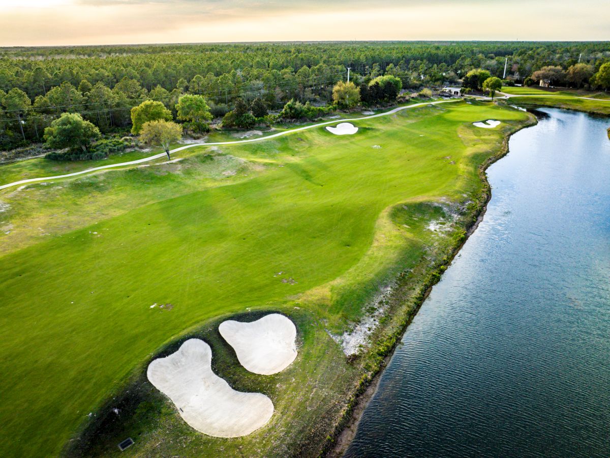 Aerial view of a golf course with green fairways, sand bunkers, and a water hazard on the right. Trees surround the distant background.