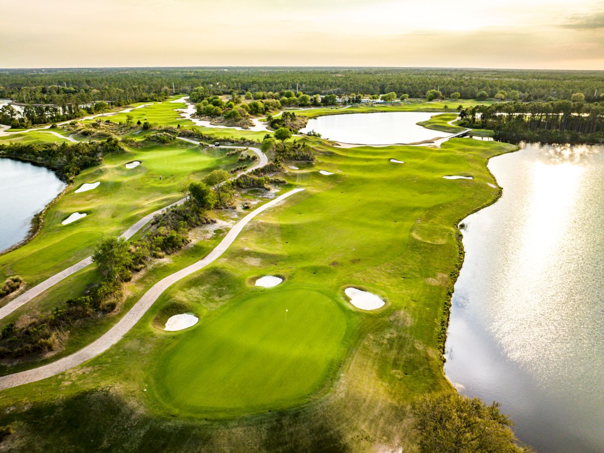 Aerial view of a scenic golf course with lush greens, sand bunkers, water hazards, and winding paths surrounded by trees and a lake.