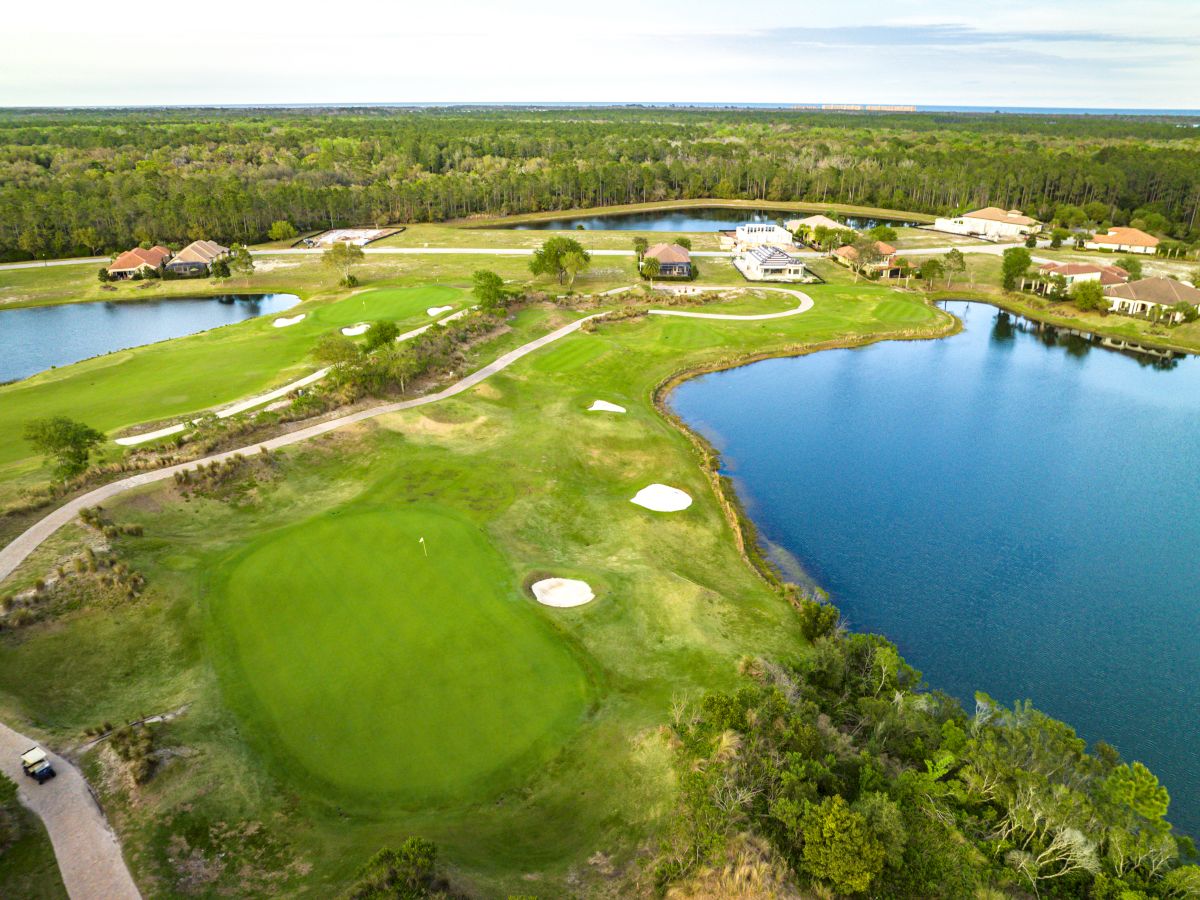 An aerial view of a golf course with a lake, sand traps, greenery, and several buildings in the background under a partly cloudy sky.