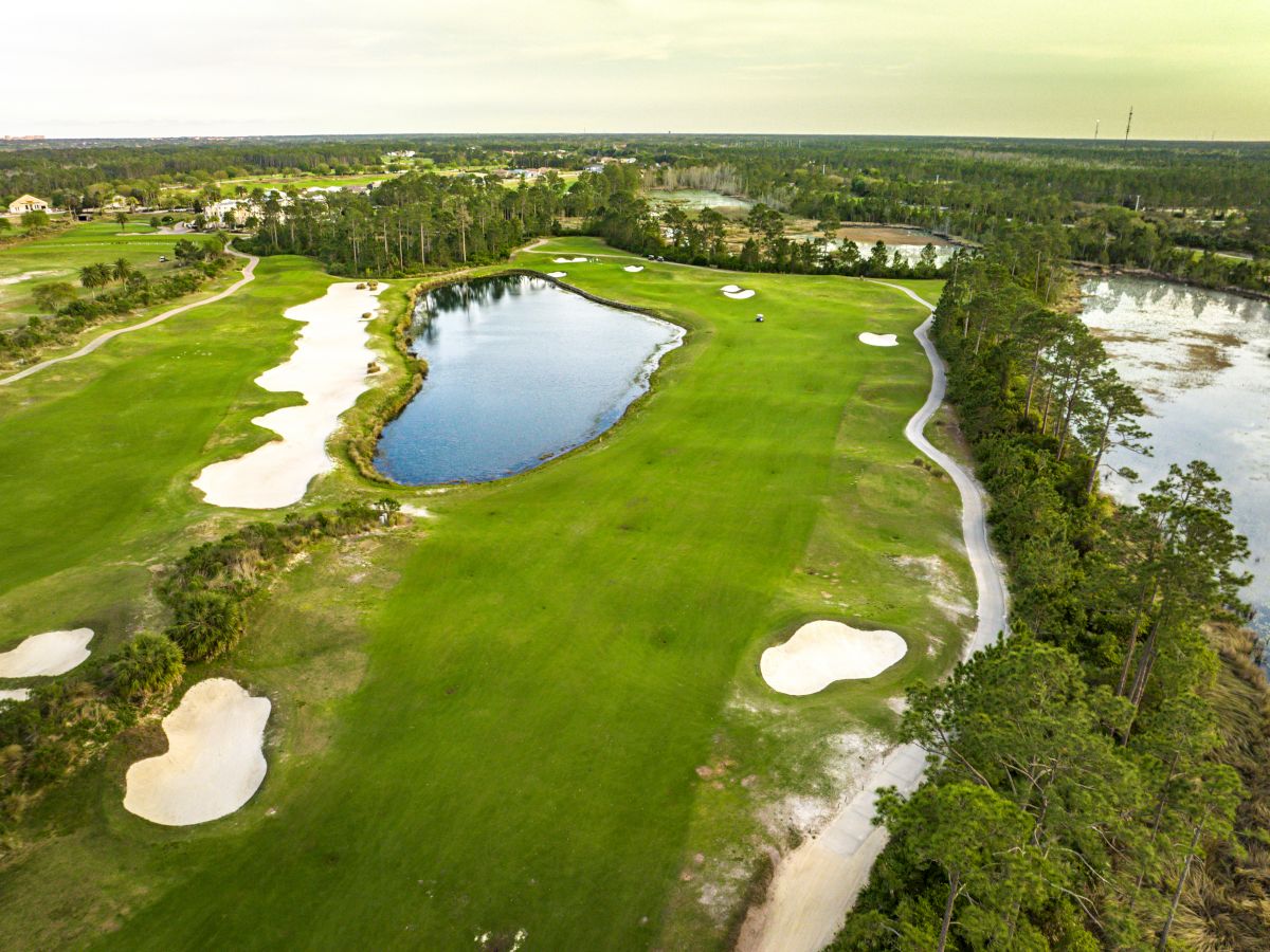 Aerial view of a golf course with sand bunkers, a water hazard, and lush greenery, bordered by trees and open skies.