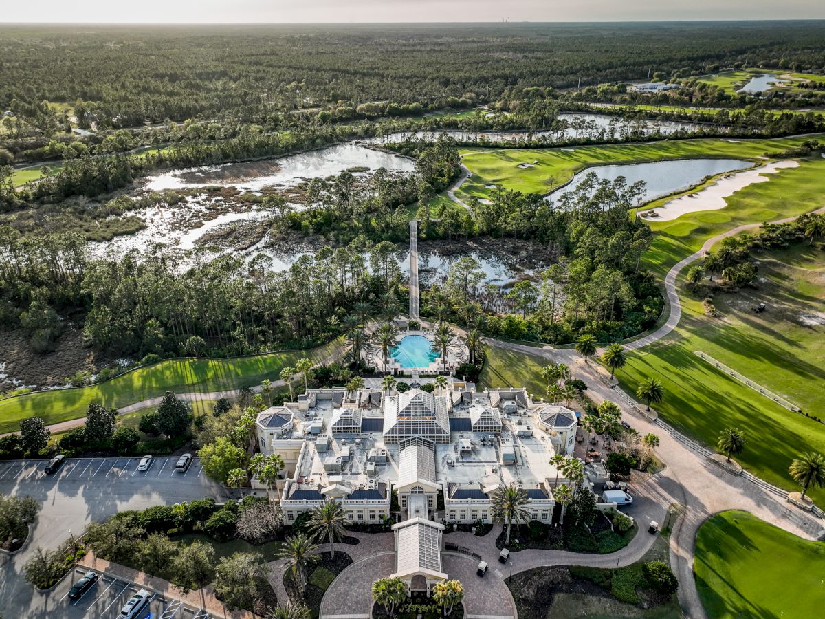 An aerial view of a large building surrounded by lush greenery, a circular pool, and a golf course, with a forested area in the background.